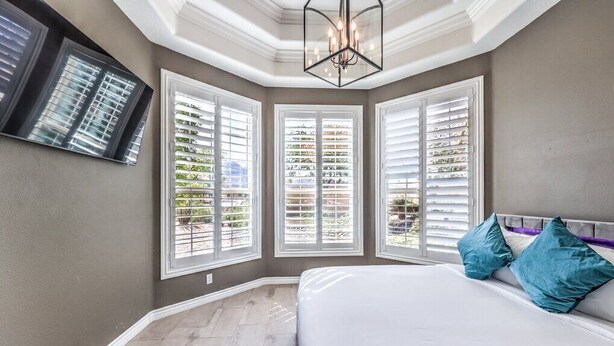 A bedroom with a large bed, a chandelier, and three windows with blinds.