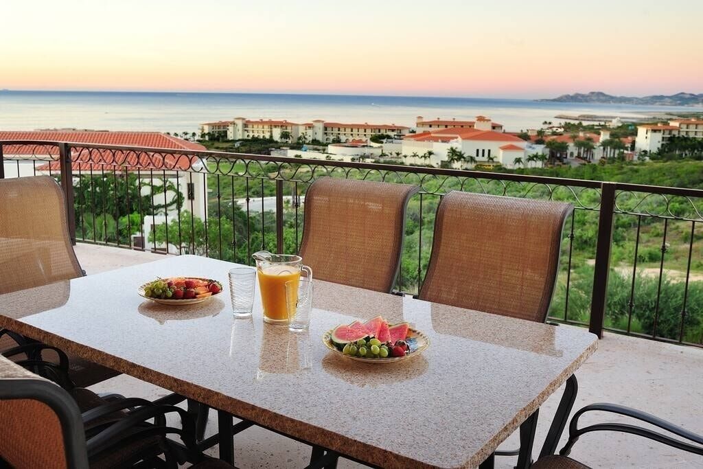 Una mesa con alimentos para el desayuno y un vaso de jugo de naranja en un balcón con vistas al mar.