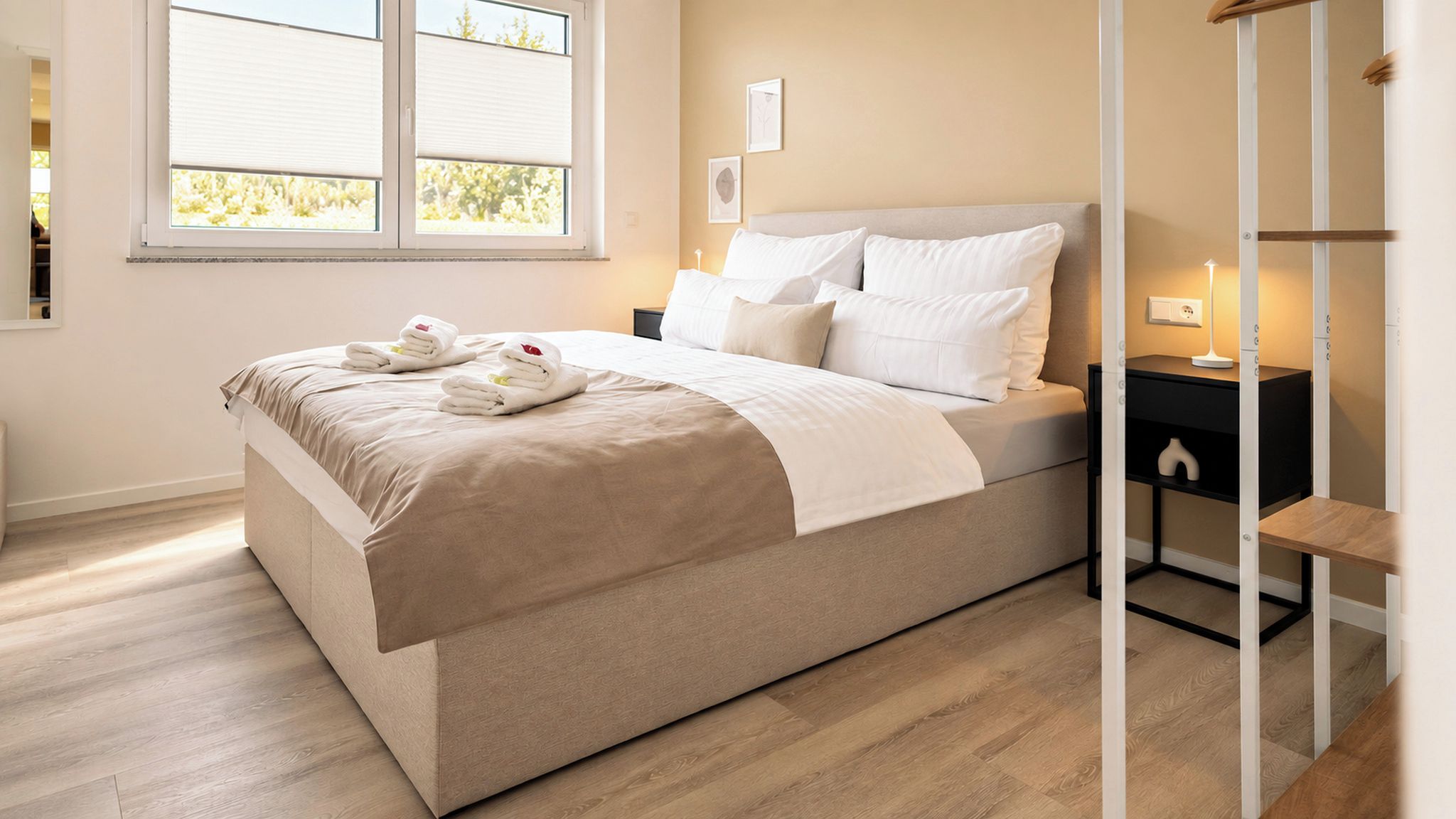 A neatly made bed with a beige bedspread and pillows, a bedside table with a lamp, and a window with blinds.
