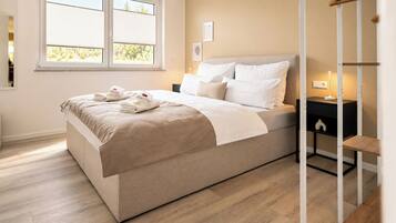 A neatly made bed with a beige bedspread and pillows, a bedside table with a lamp, and a window with blinds.
