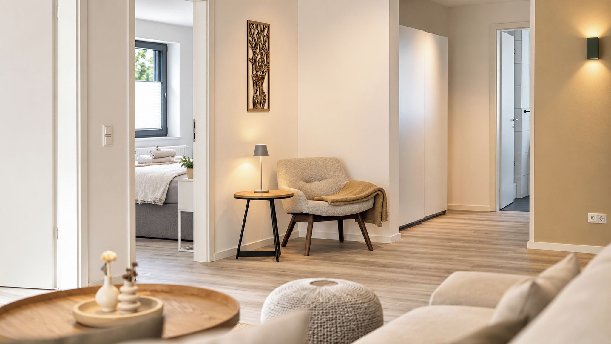 A modern living room with a sofa, a side table, a lamp, and a chair. There is a hallway leading to a bedroom with a bed and a window.