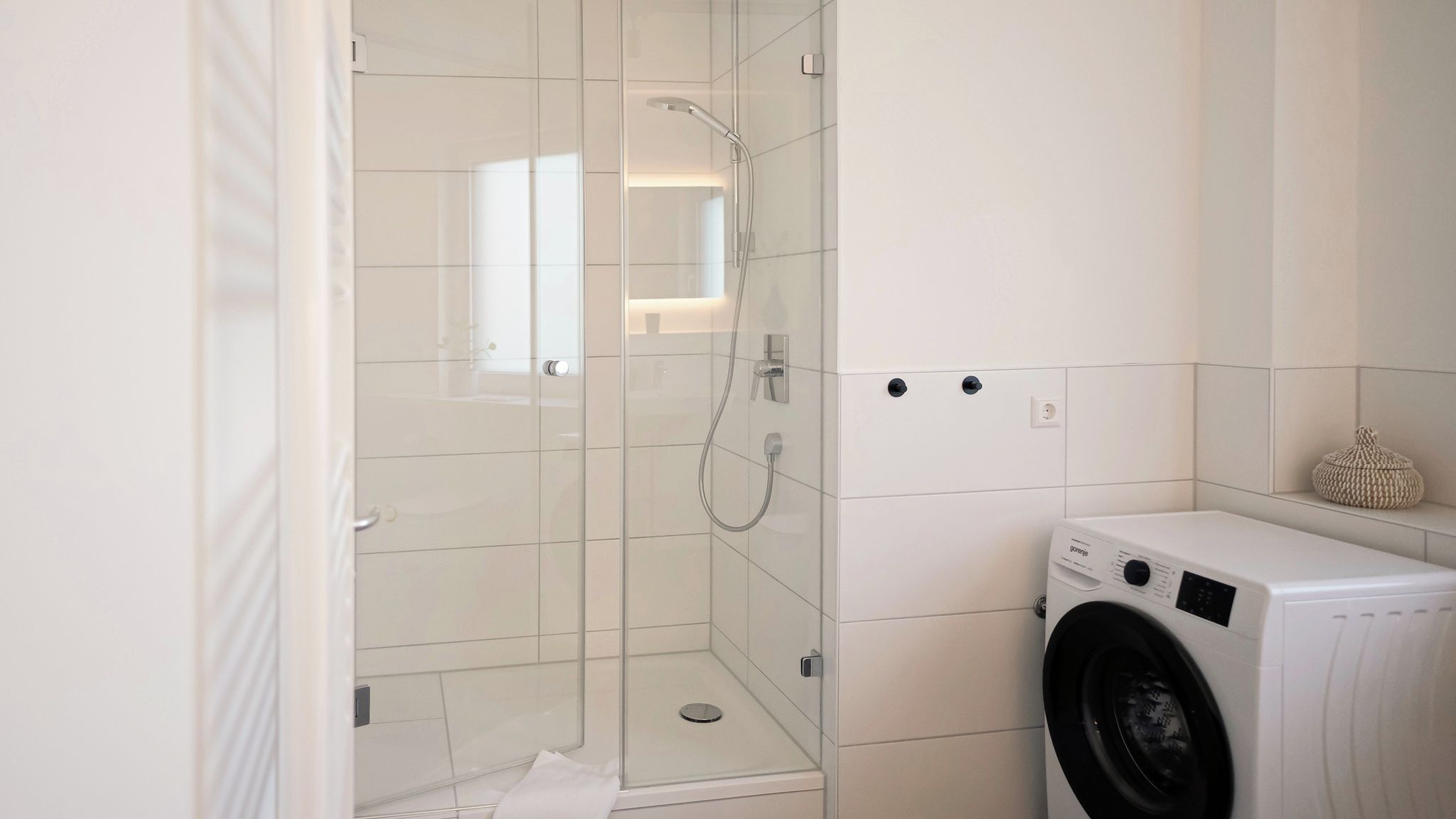 A bathroom with a glass-enclosed shower, a washing machine, and a small shelf with a decorative item.