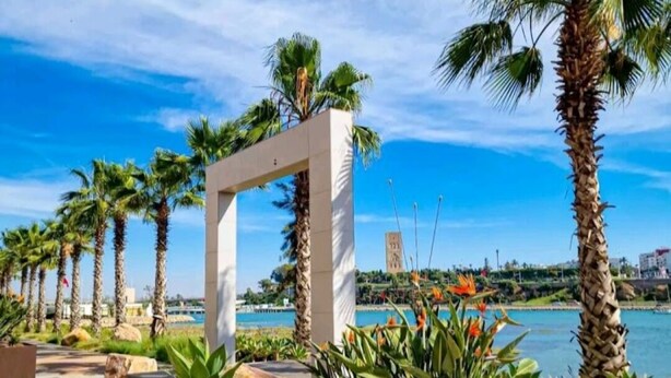 Un paseo marítimo con palmeras, una estructura de arco blanco y vistas al mar y edificios al fondo.