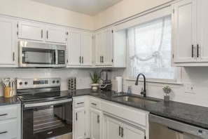 A modern kitchen with white cabinets, stainless steel appliances, a built-in oven, and a window with blinds.
