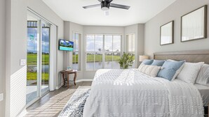 A bedroom with a large bed, a ceiling fan, a TV, and a view of a green landscape through the sliding glass door. - Rare NEW 2/2 Condo in Naples with Lake & Golf View! Home Away from Home (Naples)