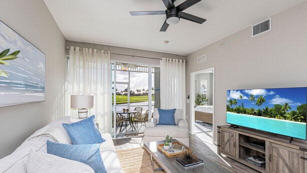 A modern living room with a sofa, coffee table, TV, and a view of a tropical beach through large windows. - Rare NEW 2/2 Condo in Naples with Lake & Golf View! Home Away from Home (Naples)