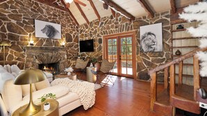 A rustic interior with stone walls, a fireplace, a wooden staircase, and a cozy seating area with a sofa and armchairs.