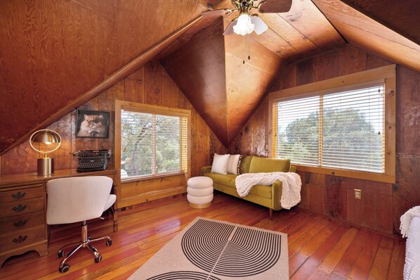 A cozy attic bedroom with wooden walls, a desk with a lamp, a green sofa, and large windows.