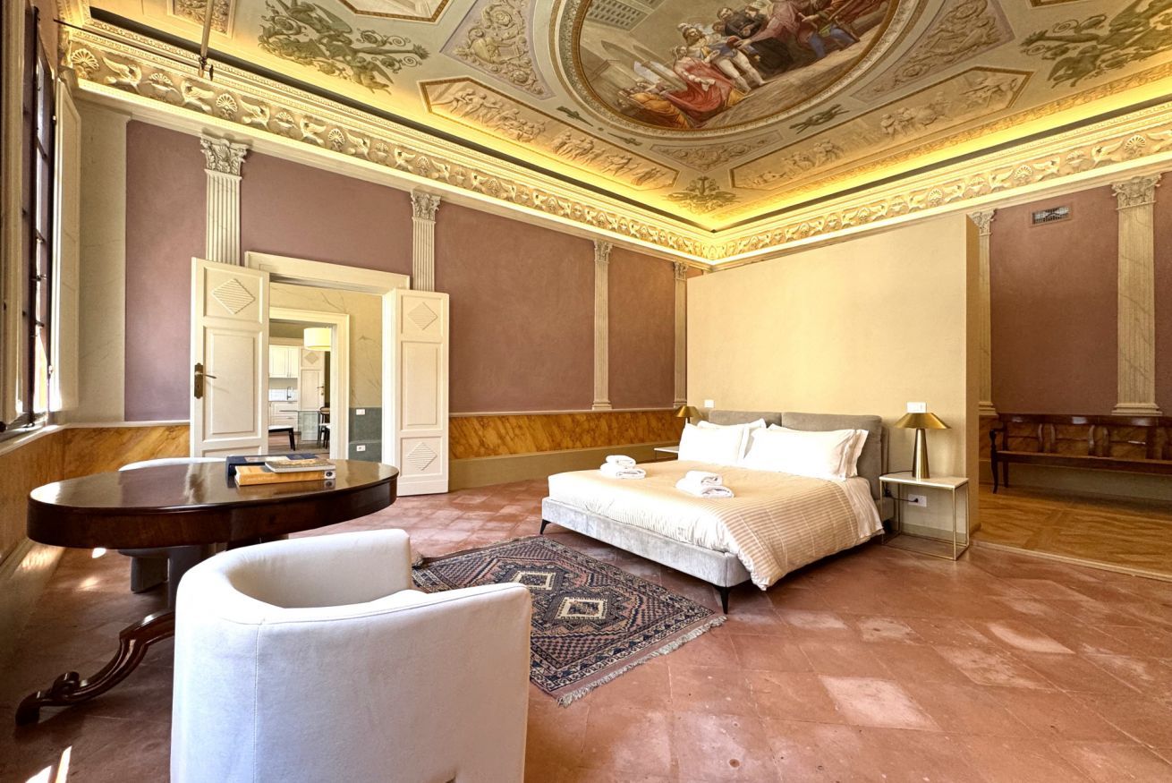 Amplia habitación de hotel con una cama grande, una mesa redonda, un sillón blanco y un techo decorativo.