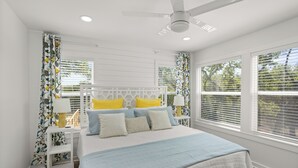 A bedroom with a white bed, multiple pillows, a nightstand with a lamp, and a ceiling fan.