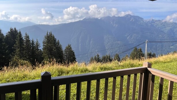 A mountain view from a wooden deck with a railing, overlooking a forested area and distant mountains.