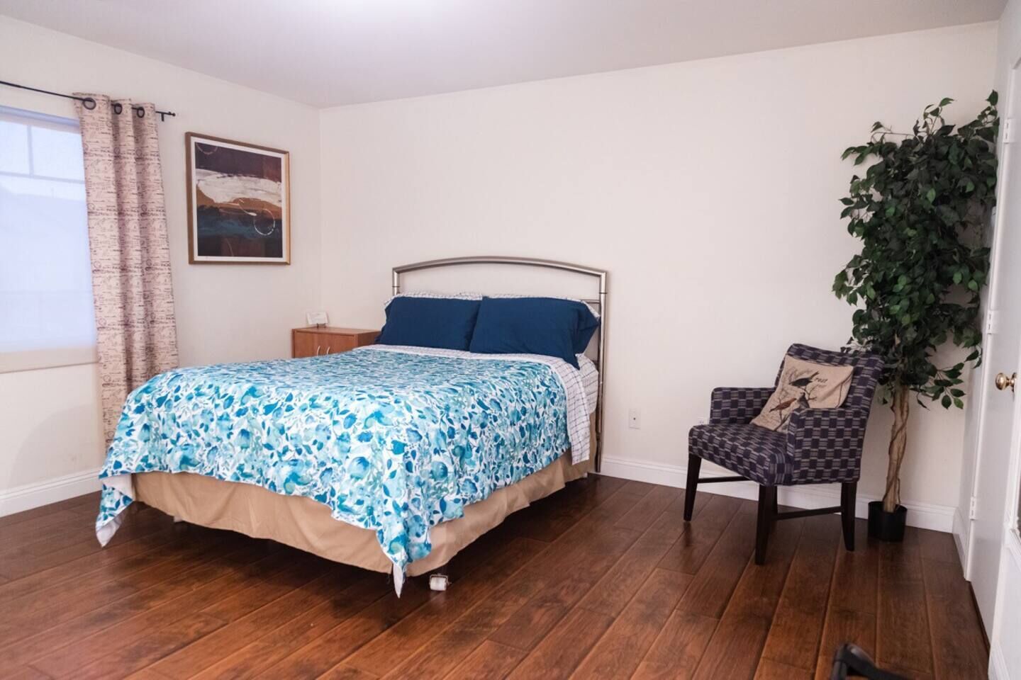 Un dormitorio con una cama, una silla, una planta y un cuadro en la pared.