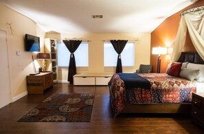 Une chambre à coucher comprenant un lit, des tables de chevet, un bureau, une chaise, un téléviseur et deux fenêtres avec des rideaux.