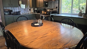 Una mesa de comedor de madera con sillas, una cocina con armarios verdes, un ventanal con vistas a árboles y una encimera con un hervidor y otros electrodomésticos.