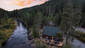 A cabin with a deck and outdoor seating, surrounded by dense forest and a river. - Riversong Lodge by Avantstay Sauna + Hot Tub On The Truckee River (Truckee)
