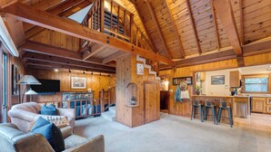 A spacious living area with a high ceiling, wooden beams, a sofa with cushions, a coffee table, and a kitchen area with a bar and stools. - Riversong Lodge by Avantstay Sauna + Hot Tub On The Truckee River (Truckee)