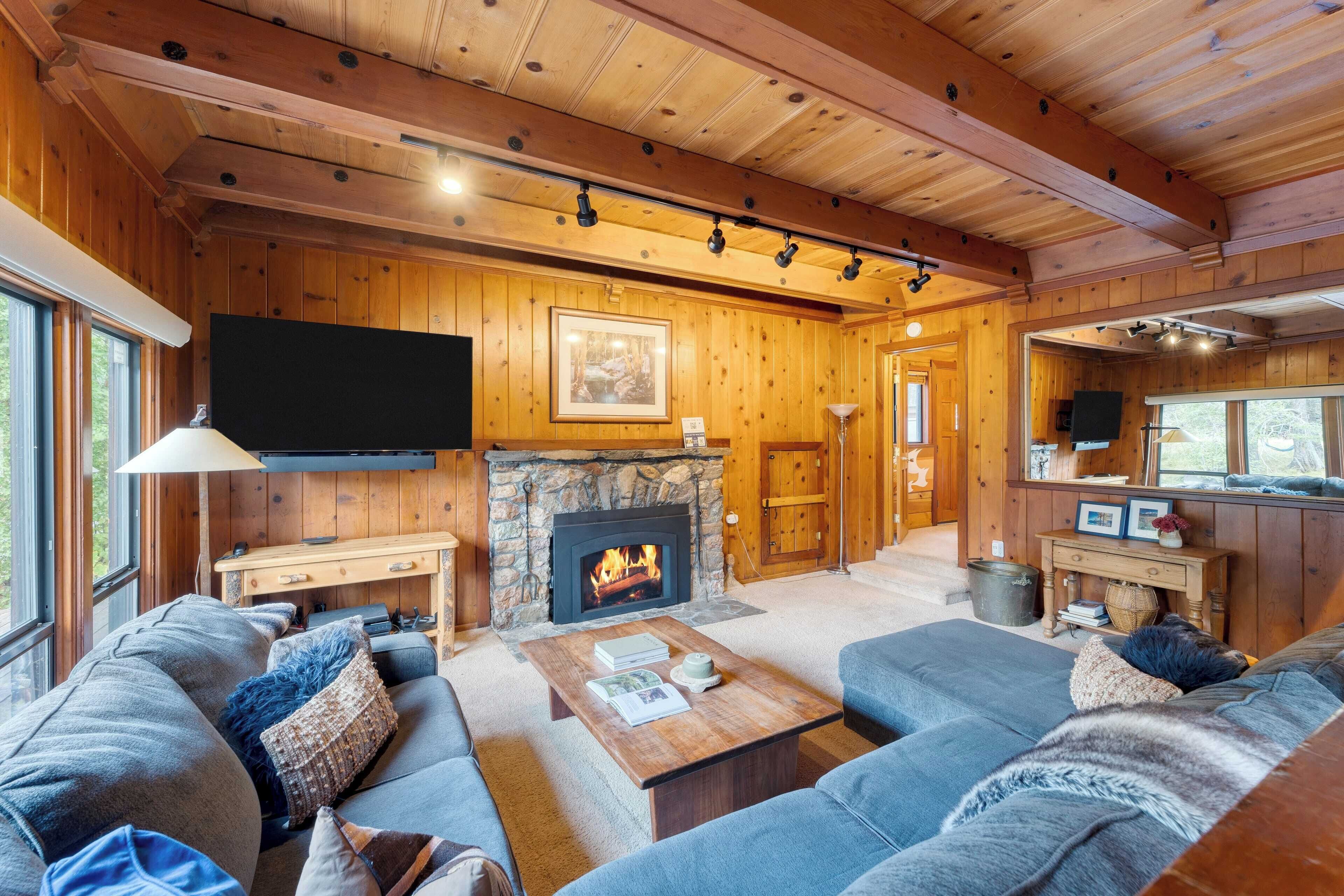 A cozy log cabin interior with a stone fireplace, wooden walls, a large sofa, and a flat-screen TV mounted on the wall.