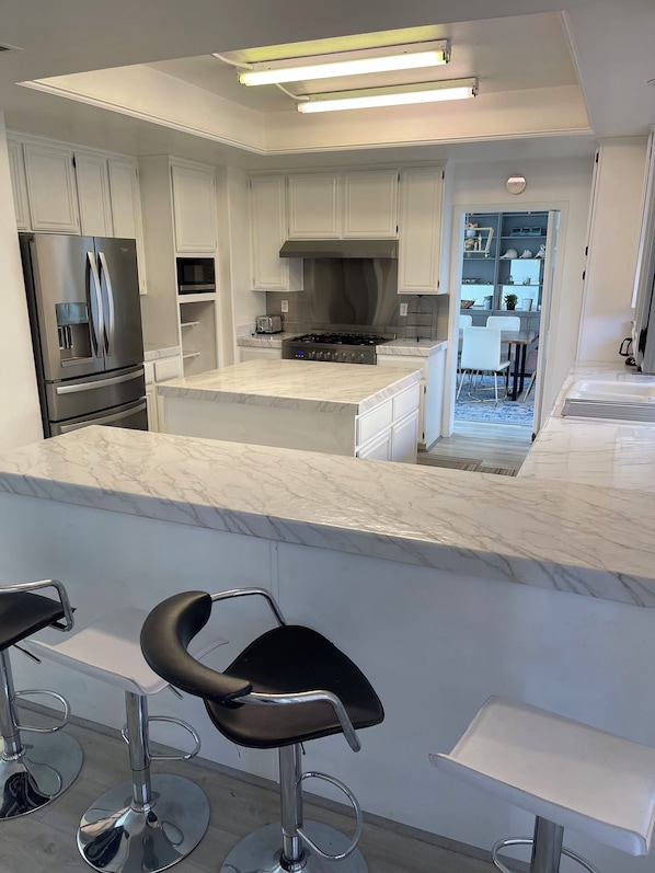 A modern kitchen with white cabinets, stainless steel appliances, and a marble countertop. There are two bar stools with black seats.