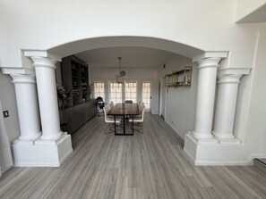 A dining area with a large table and chairs, flanked by white columns and a wooden floor.