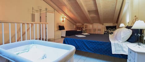 A cozy attic bedroom with a crib, a bed with a blue bedspread, and a bedside table with a lamp.