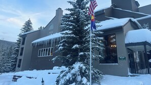 A building with a snow-covered roof, a flagpole with an American flag, and a sign indicating the address. - 1st floor ski in & out resort condo with hot tubs, fitness room, covered garage. (Winter Park)