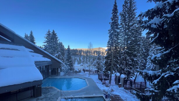 A heated outdoor pool surrounded by snow-covered trees and a building with a snow-covered roof. - 1st floor ski in & out resort condo with hot tubs, fitness room, covered garage. (Winter Park)