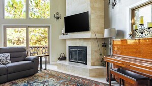 A living room with a fireplace, a sofa, a bench, and a television mounted above the fireplace. - Mystic Ridge by Avantstay Game Room + Views (Lake Arrowhead)