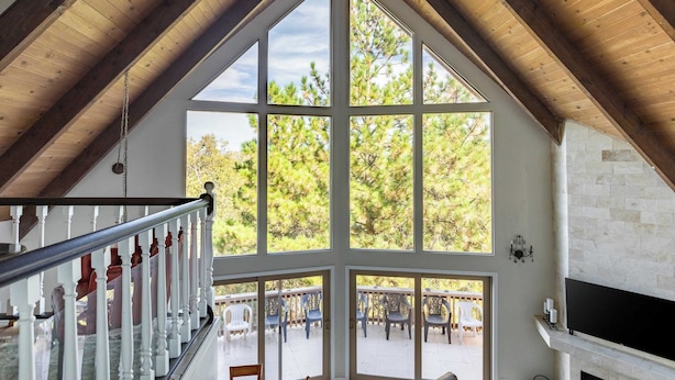 A spacious room with a large window, wooden ceiling beams, a staircase, and a fireplace. - Mystic Ridge by Avantstay Game Room + Views (Lake Arrowhead)