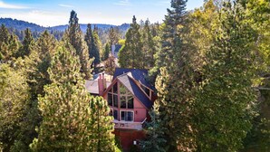 A cabin surrounded by dense forest, with a prominent chimney and large windows. - Mystic Ridge by Avantstay Game Room + Views (Lake Arrowhead)