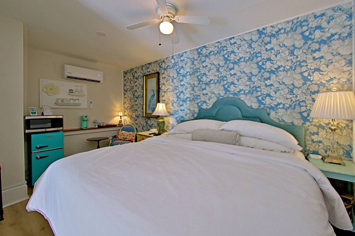 Un dormitorio con una cama grande, un ventilador de techo, un microondas y una pequeña mesa con una silla.
