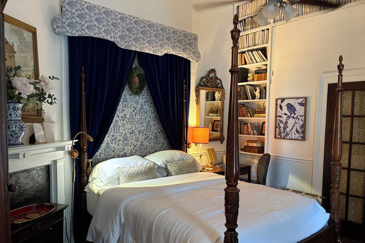 Una cama con dosel con una colcha blanca, una estantería con libros y objetos decorativos, una chimenea con un jarrón de flores, y una cortina azul con estampado floral.