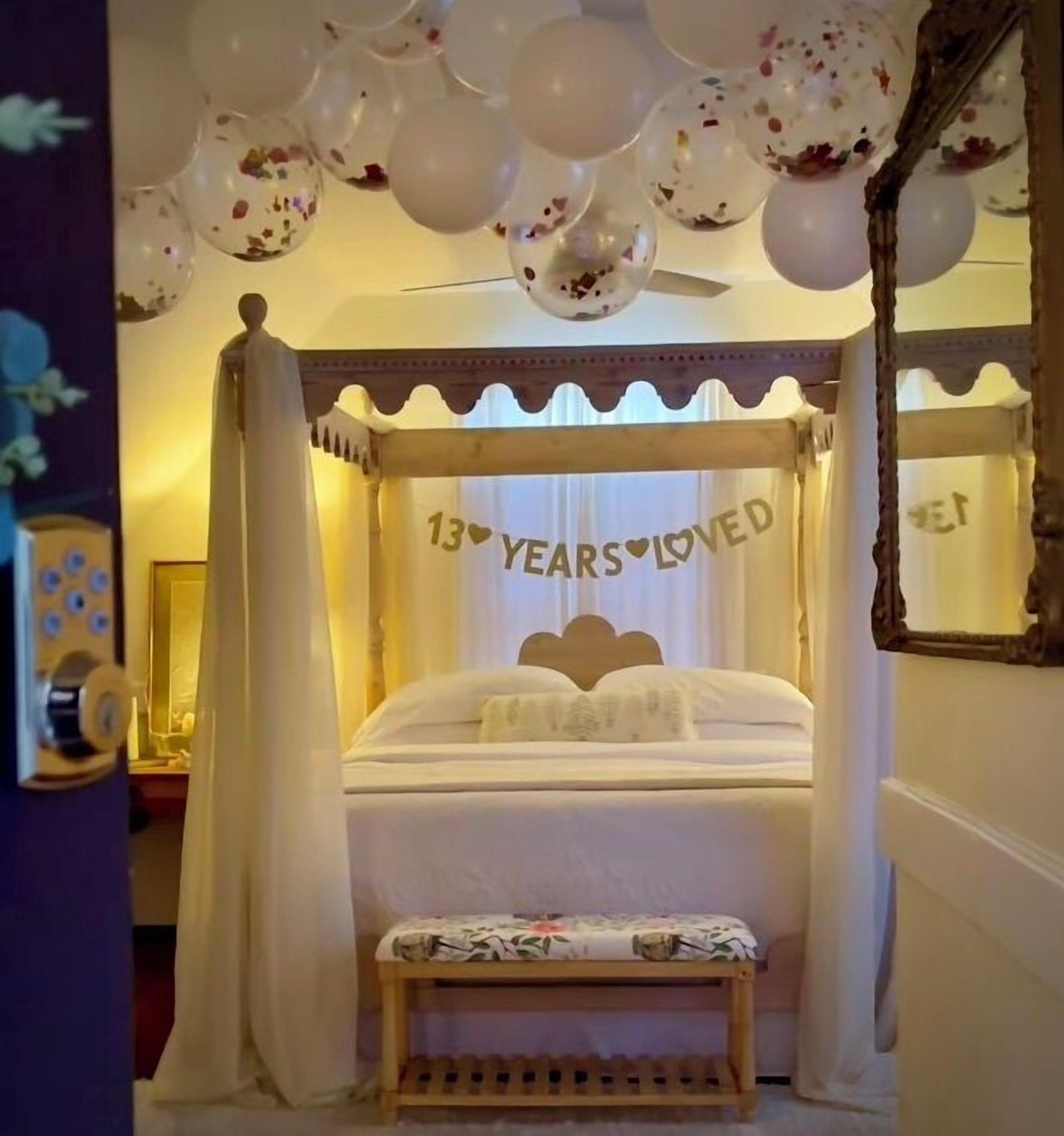 A bedroom with a canopy bed, a bedside table, and a bench. The bed has a canopy with the text "13 YEARS LOVED" on it.