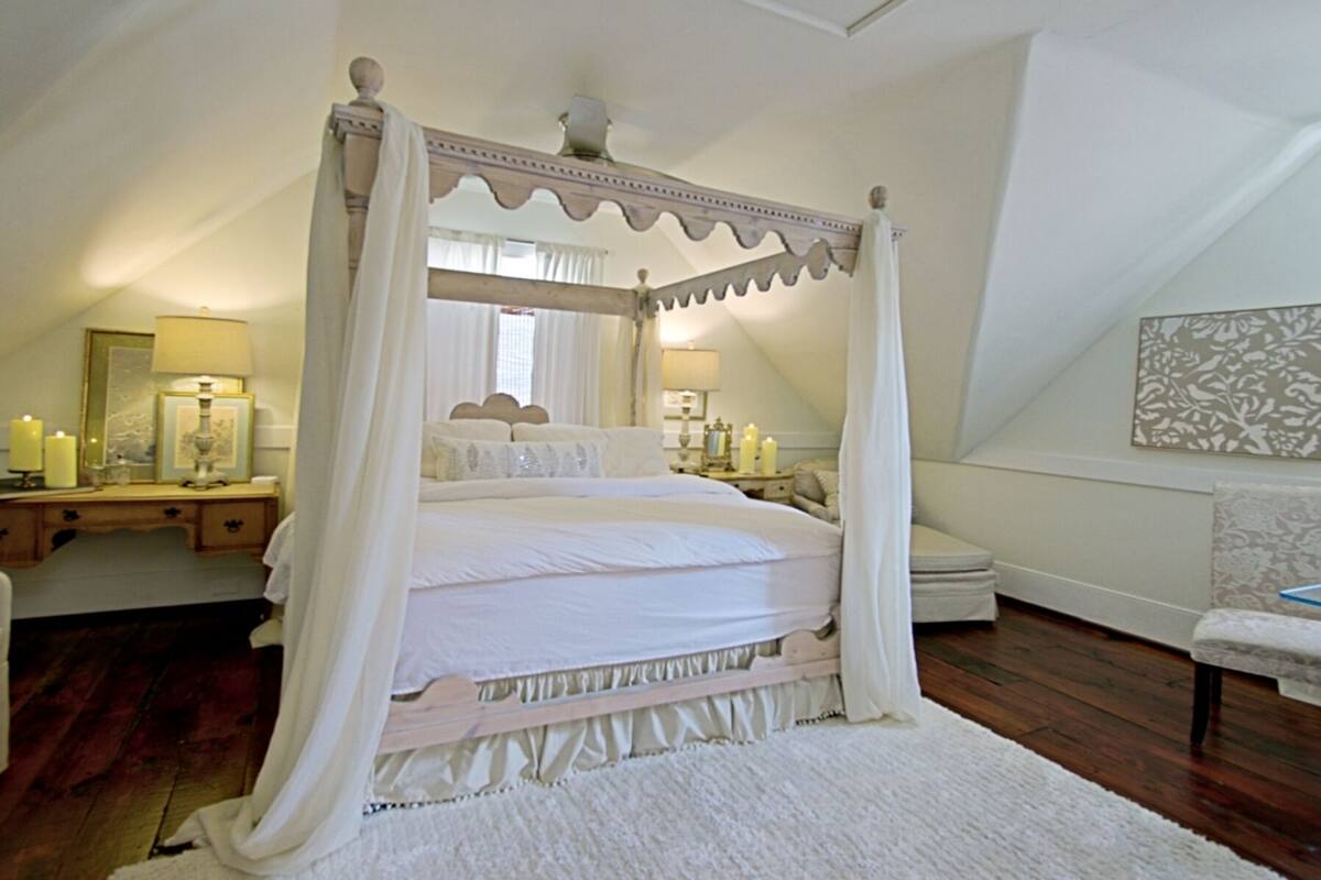 Una cama con dosel en un dormitorio con piso de madera, una mesita de noche con lámpara y una silla.