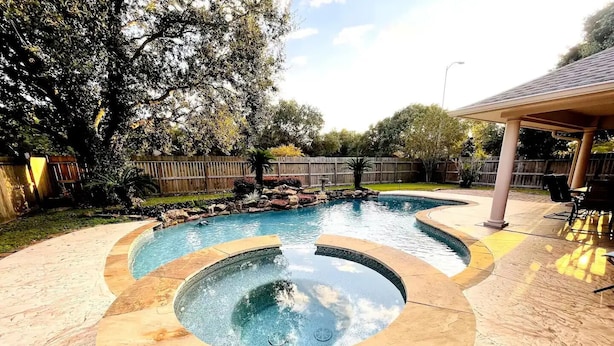 Un patio trasero con un jacuzzi circular y una piscina rectangular, rodeado por una valla de madera y árboles.