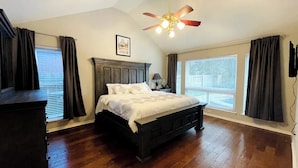 Un dormitorio con cama de madera, ventilador de techo y ventanas amplias con cortinas.