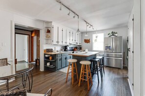 Una cocina moderna con armarios blancos, una isla central con taburetes, electrodomésticos de acero inoxidable y una zona de comedor con una mesa de vidrio y sillas de metal.