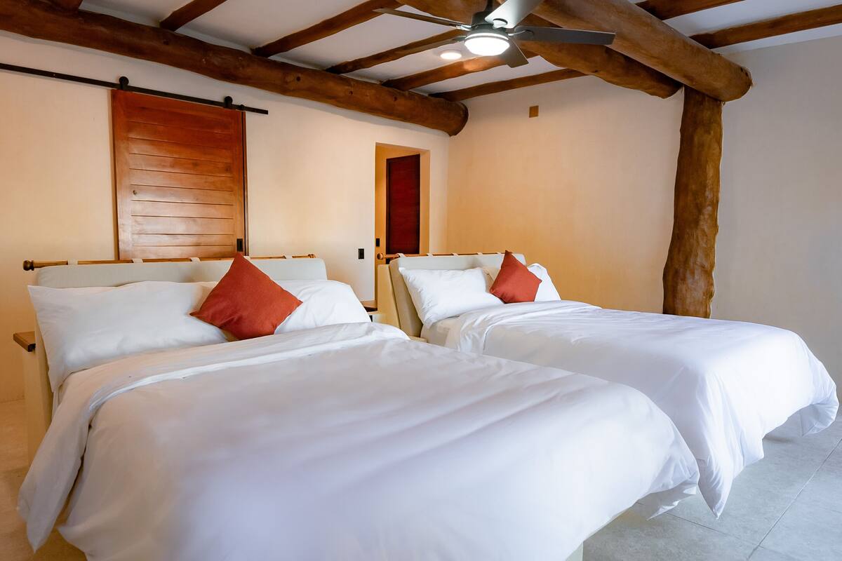 Duas camas de solteiro com roupa de cama branca e almofadas vermelhas em um quarto com vigas de madeira expostas e ventilador de teto.