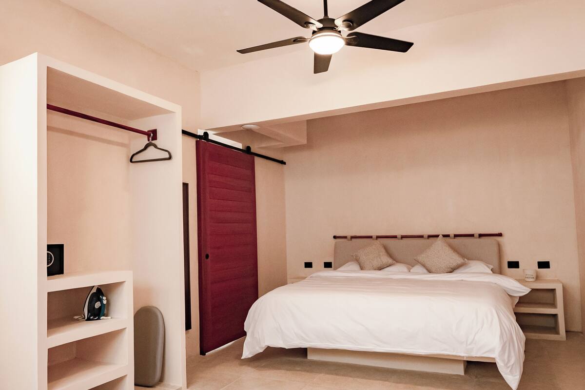 Quarto com cama, guarda-roupa, ventoinha no teto e mesa de cabeceira.