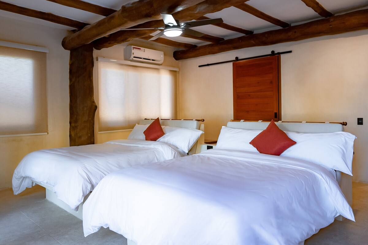 Quarto com duas camas, vigas de madeira, ventoinha no teto e ar condicionado.