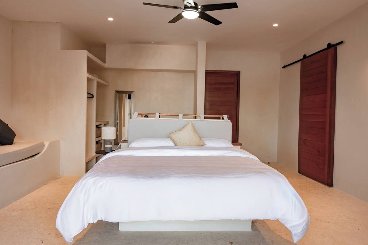 Quarto com cama, ventoinha no teto, mesa de cabeceira e porta de madeira.