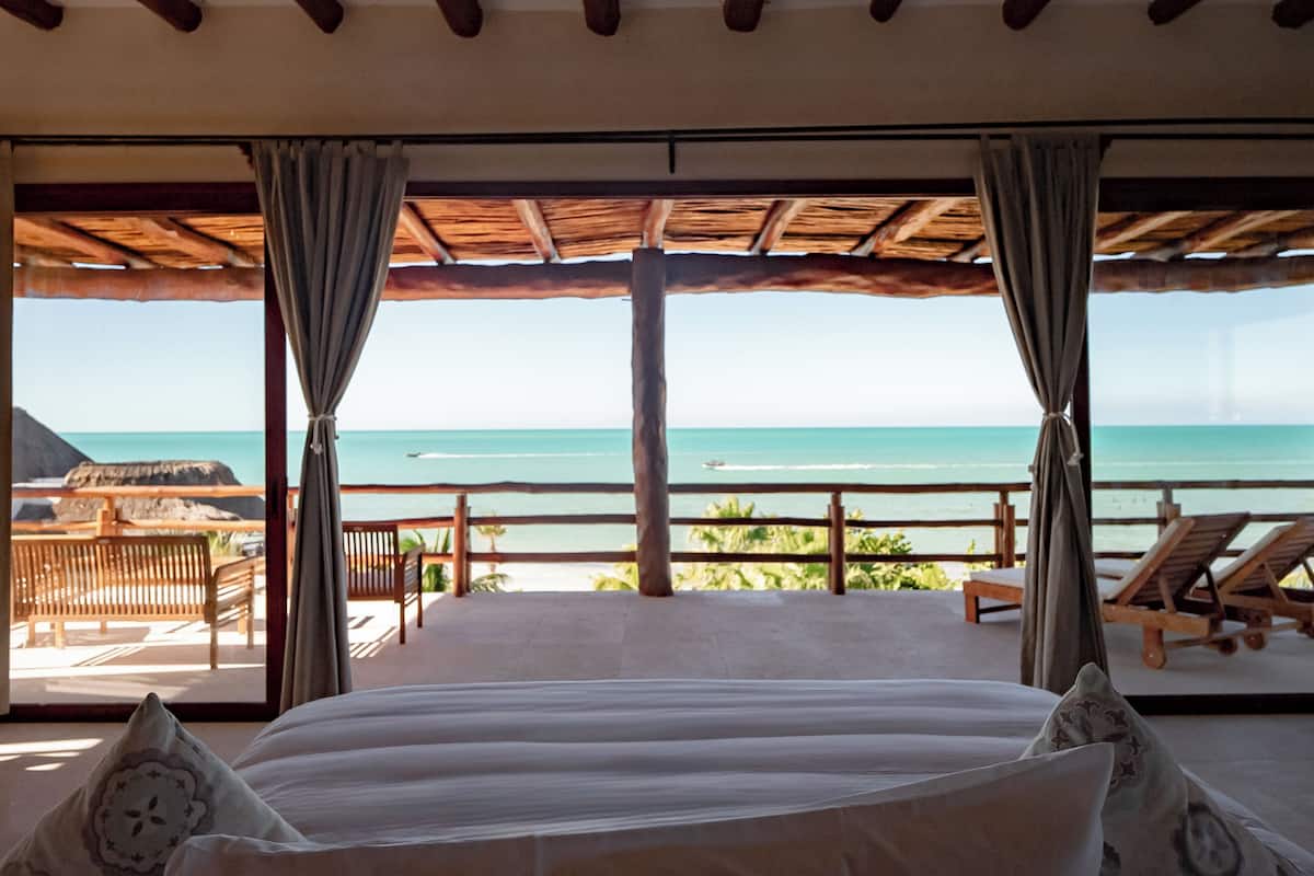 Quarto com cama grande, vigas de madeira no teto e vista para o mar.