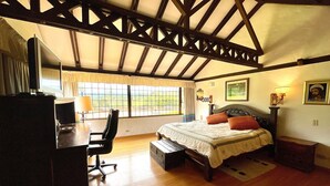 Une chambre avec un grand lit, un bureau et une chaise. Il y a une fenêtre avec des rideaux, une lampe et un tableau encadré au mur.