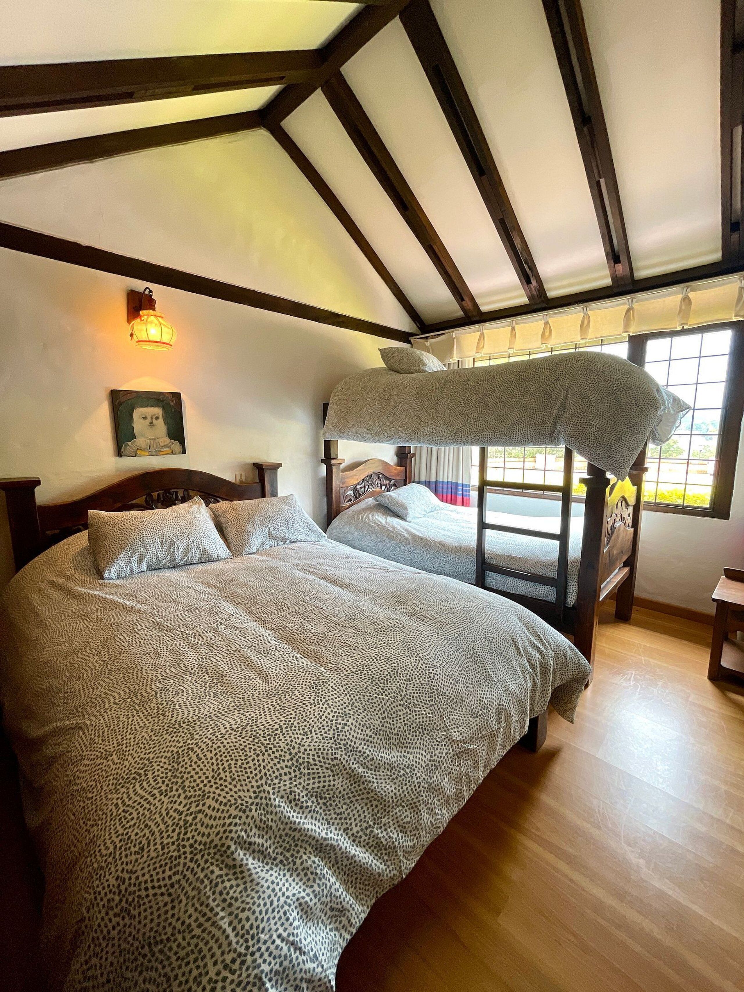 Une chambre avec deux lits, des poutres en bois au plafond, une lampe fixée au mur et une fenêtre avec des rideaux.