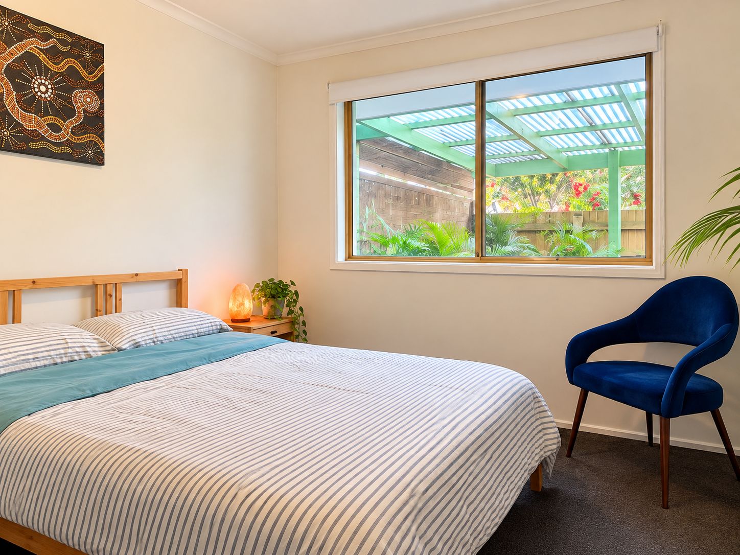 A bedroom with a bed, a chair, a window with a view of a garden, and a wall art piece.
