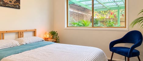 A bedroom with a bed, a chair, a window with a view of a garden, and a wall art piece.