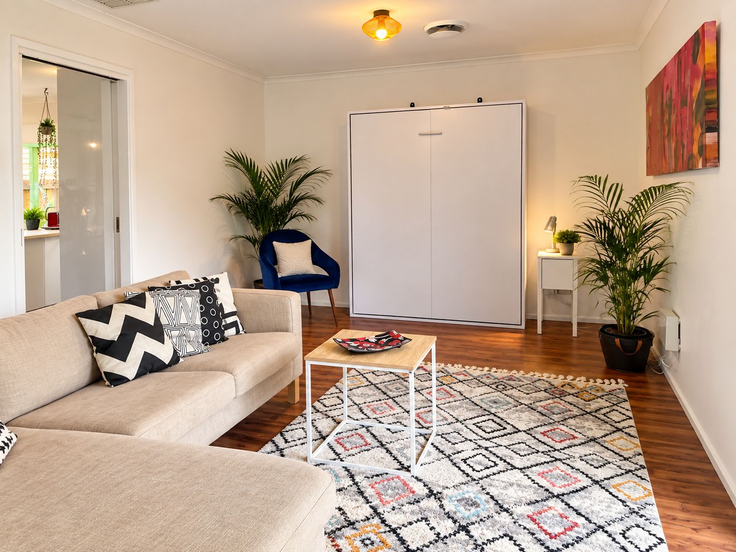 Ein Wohnzimmer mit einer Couch, einem Couchtisch und einem gemusterten Teppich. Dort ist eine Tür, ein Sessel und ein Blumentopf.