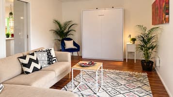 Ein Wohnzimmer mit einer Couch, einem Couchtisch und einem gemusterten Teppich. Dort ist eine Tür, ein Sessel und ein Blumentopf.