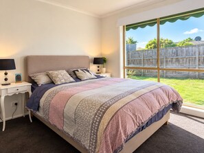 Quarto com uma cama grande, duas mesas de cabeceira, janela com vista para uma cerca de madeira e uma mesa de apoio com luminária.