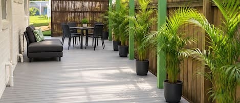 A covered patio area with a wooden deck, a seating area with a sofa and chairs, and potted plants.