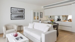 A modern living room with a white sofa, a small coffee table with a vase, and a dining area with a table and chairs. There are framed pictures on the wall and a kitchen area with cabinets and appliances.